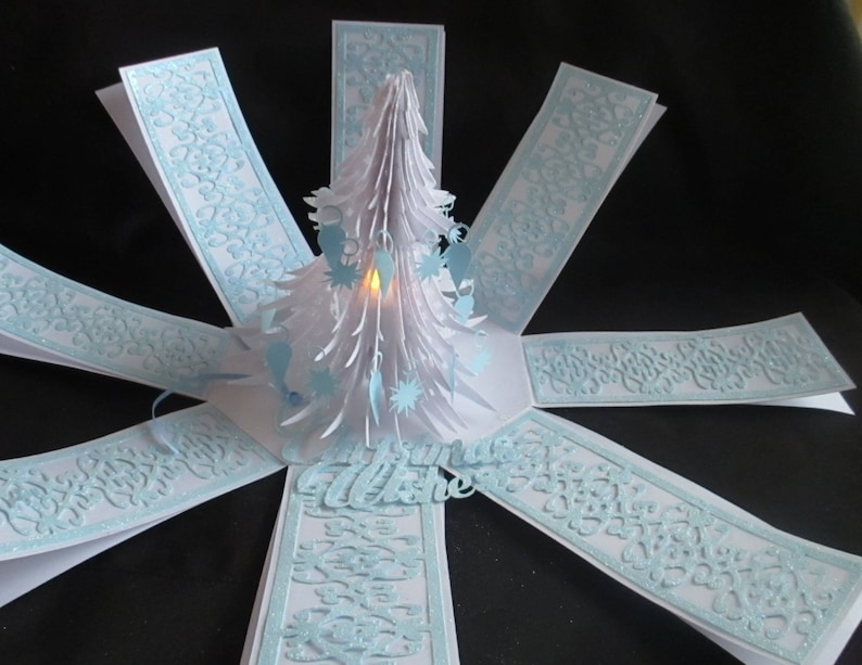 May include: A white paper Christmas tree pop-up card with a light blue patterned design. The card is open and the tree is illuminated with a small light.