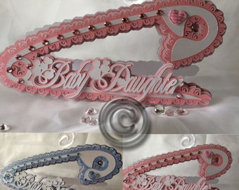 Puede incluir: Un broche de seguridad para bebé de color rosa y blanco con las palabras "Baby Daughter" en letras blancas. El broche está decorado con diamantes de imitación y un pequeño corazón rosa.
