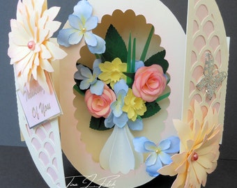 May include: A 3D pop-up card with a bouquet of flowers in a vase. The card is decorated with a scalloped edge and a delicate floral pattern. The card has a tag that says "Thinking of You".
