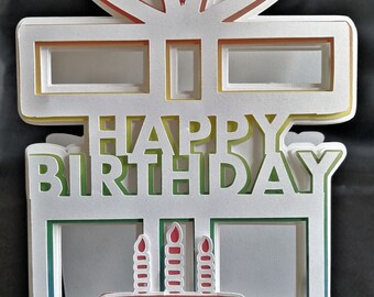 May include: A white papercraft gift box with a rainbow layered design. The box has a bow on top and says "Happy Birthday" on the front. Inside the box is a small cake with three candles.