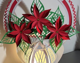 Op de afbeelding: Een witte papieren vaas met een lichtje erin, versierd met drie rode kerststerren en groene bladeren. De vaas heeft een decoratief ontwerp en de woorden "Merry Christmas" staan in een cirkel rond de bovenkant van de vaas.