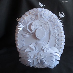 Può includere: Scultura in carta bianca di una scimmia circondata da foglie e fiori. La scimmia è realizzata in carta a strati e ha un effetto 3D. La scultura è decorata con perline bianche attorno al bordo.