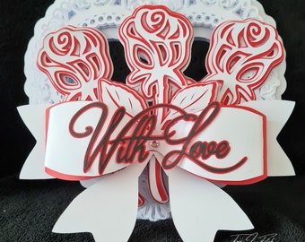 May include: A white heart-shaped card with a red and white bow and three red roses with white centers. The card has the words "With Love" written in black on the bow.