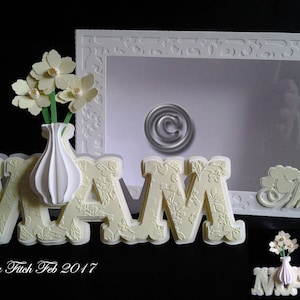 May include: A white frame with a decorative border and a clear window. The frame is decorated with the word "Mom" in white letters. A white vase with yellow flowers is in front of the frame. The vase is decorated with the word "Mom" in yellow letters.