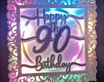 May include: A white card with a decorative frame and a colorful light shining through the cutouts. The card says "Happy 90th Birthday" in black lettering.