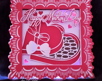 May include: A red and white layered Mother's Day decoration. The design features a heart-shaped box of chocolates with a bow, surrounded by a decorative frame. The words "Happy Mother's Day" are written across the top.