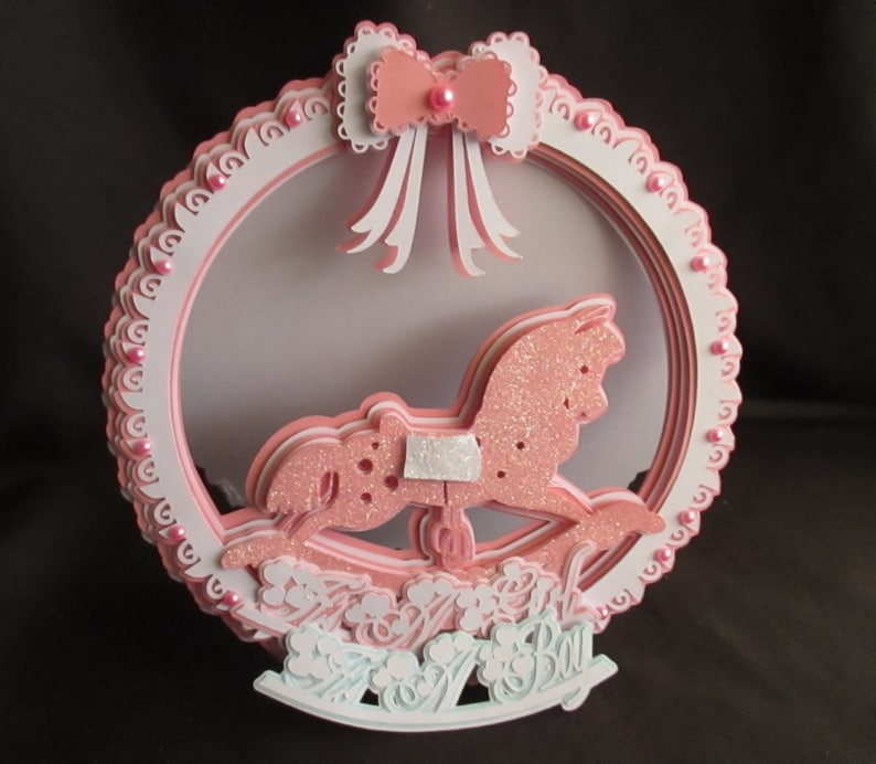 May include: A pink and white layered card with a pink glitter rocking horse and the words "It's a Boy" in a white and green font.