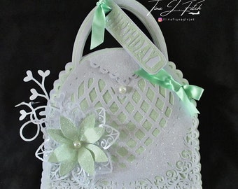 Puede incluir: Un bolso de papel blanco y verde con un diseño de encaje y un lazo verde. El bolso tiene una etiqueta que dice "Cumpleaños".