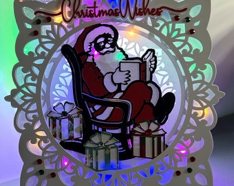 Op de afbeelding: Witte houten kerstkaart met een uitgesneden ontwerp met Sinterklaas die een boek leest in een schommelstoel. De kaart is verlicht met kleurrijke LED-lampjes en heeft de tekst "Christmas Wishes" bovenaan.