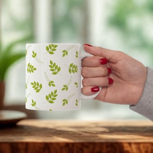 May include: White ceramic mug featuring a repeating pattern of green leaves and the text "Fresh Forest". The mug has a white handle. The mug is held by a person with red nail polish. The mug is on a wooden surface.