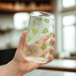 Puede incluir: Un vaso transparente en forma de lata con un diseño de hojas verdes. El vaso se sostiene en una mano y el diseño incluye las palabras "Fresh Spring". El vaso está diseñado para bebidas.