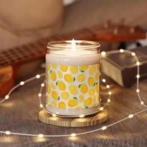 May include: A lit candle in a glass jar with a yellow lemon pattern. The candle sits on a small wooden coaster, surrounded by a string of lights. A guitar and a book are in the background.