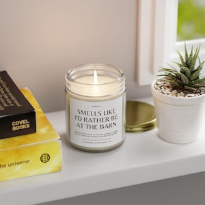 May include: A lit, clear glass jar candle with a white label that reads "Smells Like I'd Rather Be at the Barn." A small succulent in a white pot sits nearby. A book and a gold lid are also visible.