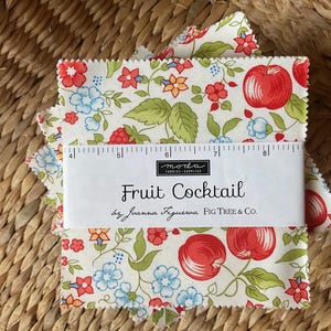 Può includere: Quadrati di tessuto con un motivo floreale e fruttato. Il tessuto presenta mele rosse, fiori blu e foglie verdi su sfondo crema. Un righello è posto sopra il tessuto e il testo "Fruit Cocktail" è visibile.