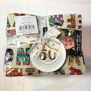 Könnte beinhalten: Ein Stoffbündel, verpackt in durchsichtigem Kunststoff. Der Stoff hat ein Vintage-Design mit Text und Bildern. Das Bündel ist mit einem Band und einem Etikett mit der Aufschrift "50 Jahre" gebunden. Das Bündel misst 45,7 cm x 55,9 cm.