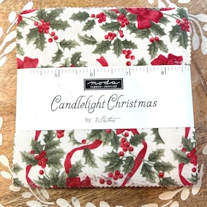 May include: A stack of square fabric pieces featuring a white background with a pattern of green holly leaves, red berries, and red ribbons. A paper band labeled "Candlelight Christmas by 3 Sisters" with a 5 to 8 inch ruler is wrapped around it.