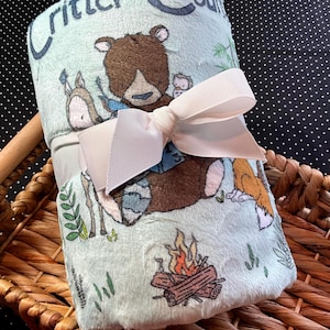May include: A rolled-up, light blue plush blanket with a woodland animal design, tied with a cream-colored ribbon. The blanket features illustrations of a bear, deer, and fox, with the words "Critter Country" printed on it.