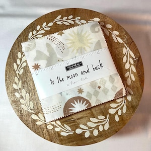 May include: Fabric squares with a celestial design, featuring stars, moons, and geometric patterns in neutral tones. The fabric is wrapped with a label that reads "to the moon and back". The fabric is on a round wooden tray with a white leaf design.
