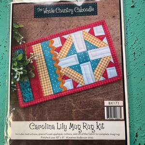 May include: A Carolina Lily Mug Rug Kit in a clear plastic package. The kit includes instructions and precut fabric. The finished mug rug size is 10" x 6". The design features a patchwork pattern with red, blue, and yellow fabrics.