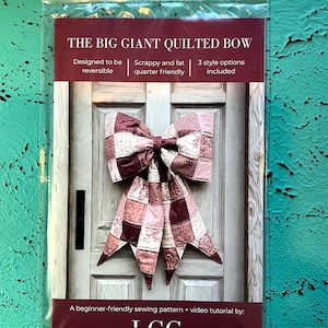 May include: A clear plastic bag contains a sewing pattern for a large quilted bow. The bow is made of patchwork fabric in shades of burgundy, pink, and cream. Text on the packaging reads "THE BIG GIANT QUILTED BOW" and "A beginner-friendly sewing pattern + video tutorial by: LGC."