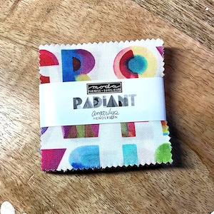 Op de afbeelding: Een stapel stofvierkanten met een kleurrijk geometrisch patroon, verpakt met een witte band. De band toont het woord "RADIANT" en de merknaam "Moda Fabrics & Supplies". De stof heeft een mix van heldere kleuren.
