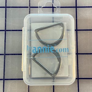 Può includere: Un contenitore di plastica trasparente con due anelli a D color argento. Gli anelli sono a forma di semicerchio con un lato piatto. Il testo "byannie.com" è visibile sulla confezione.