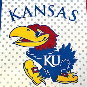May include: A white fabric with a blue border and a repeating pattern of gray polka dots. The word "KANSAS" is printed in blue above a cartoon Jayhawk mascot in red, yellow, and blue. The letters "KU" are printed on the mascot.