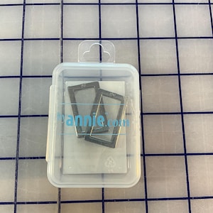 Può includere: Una custodia di plastica trasparente contenente due telai metallici rettangolari. La custodia ha un'aletta per appenderla e un logo azzurro che recita "annie.com". Lo sfondo è una griglia blu e bianca.