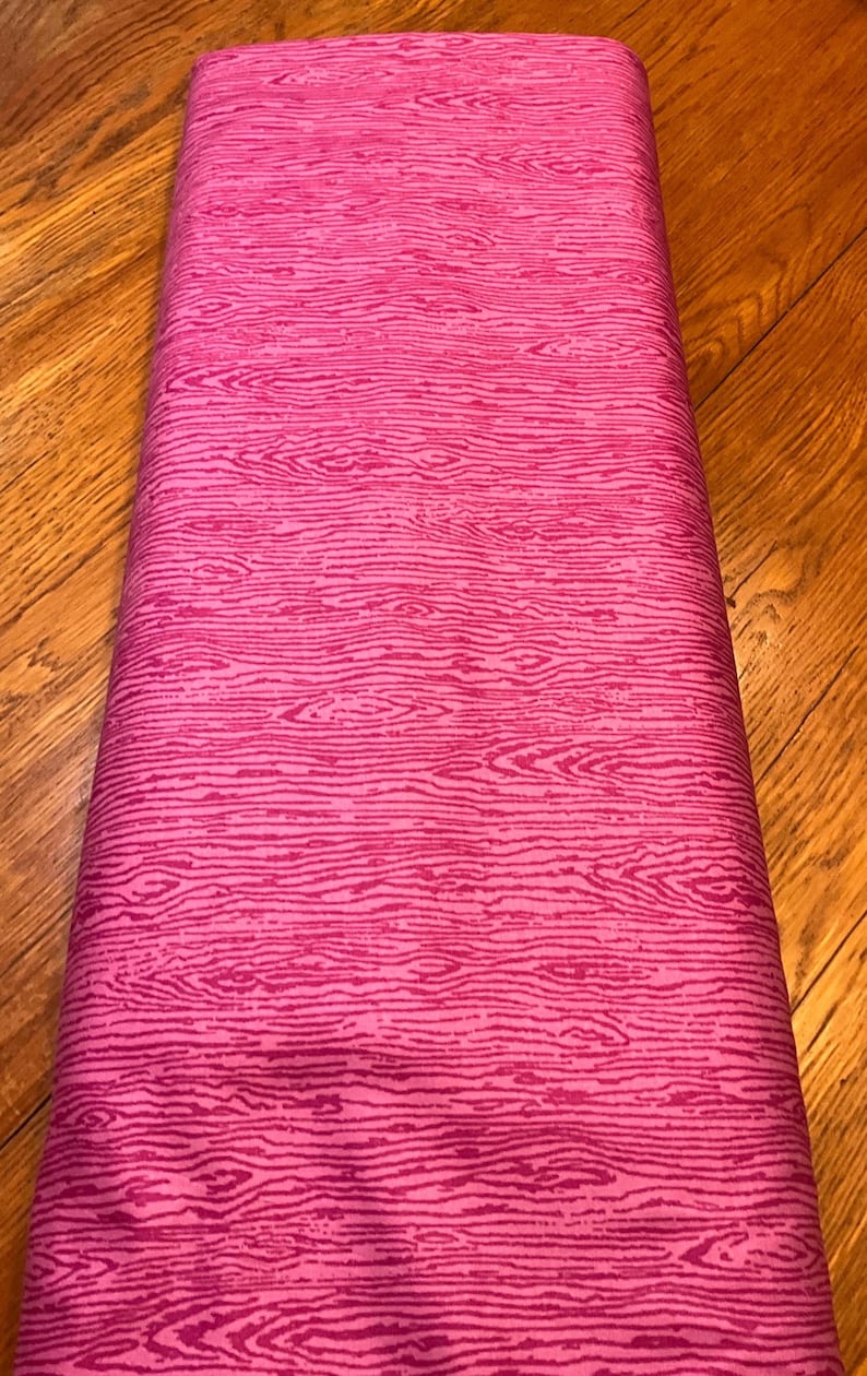 K&ouml;nnte beinhalten: Eine Rolle rosa Stoff mit Holzmaserung. Der Stoff hat einen leuchtenden Rosaton mit dunkleren rosa Linien, die die Textur von Holz nachahmen. Der Stoff ist ordentlich aufgerollt und bereit f&uuml;r N&auml;h- oder Bastelprojekte.