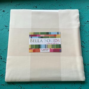 May include: A stack of cream-colored fabric squares, neatly packaged and labeled "BELLA SOLIDS moda." The label features a colorful geometric design. The fabric is arranged on a textured turquoise surface.