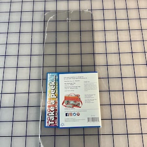 Può includere: Uno stabilizzatore di base trasparente e rettangolare con bordi arrotondati, progettato per l'uso con i modelli ByAnnie. Un'etichetta colorata con il testo "Take a peek..." e l'immagine di una borsa è attaccata.