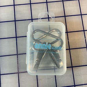 Può includere: Custodia in plastica trasparente contenente due ganci girevoli color argento. I ganci sono interbloccati e hanno una chiusura a molla. Il testo "annie.com" è stampato in blu sulla custodia.