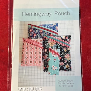 May include: A sewing pattern for a Hemingway Pouch by Center Street Quilts. The cover shows four quilted zipper pouches in various floral fabrics with diagonal zippers, presented in a clear plastic bag with printed text and grid background.