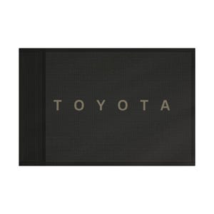 Puede incluir: Alfombrilla de coche negra rectangular con el nombre "TOYOTA" en letras grises claras. La alfombrilla tiene una superficie texturizada y un borde negro sólido.