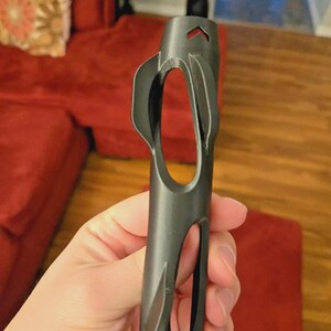 Pu&ograve; includere: Uno strumento cilindrico grigio scuro con una serie di ritagli ovali e a forma di goccia. Lo strumento ha una parte superiore appuntita con un'apertura a forma di freccia. L'oggetto &egrave; tenuto in mano, con un divano rosso sullo sfondo.