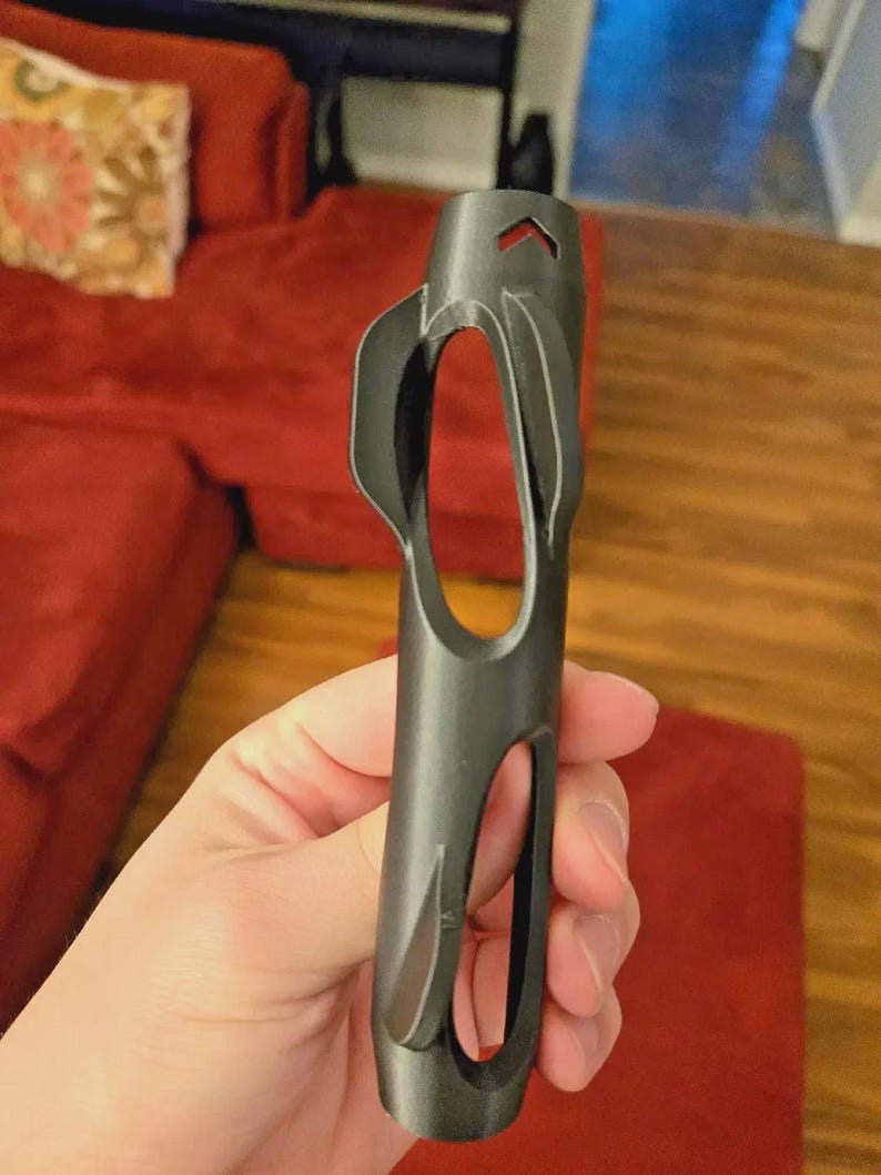 Pu&ograve; includere: Uno strumento cilindrico grigio scuro con una serie di ritagli ovali e a forma di goccia. Lo strumento ha una parte superiore appuntita con un'apertura a forma di freccia. L'oggetto &egrave; tenuto in mano, con un divano rosso sullo sfondo.