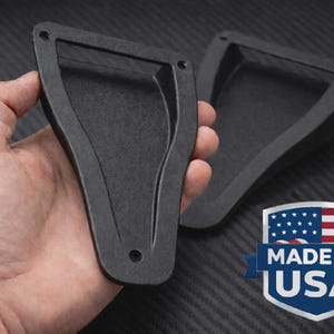 May include: Two black, angular automotive parts are displayed, one held in a hand. The parts have a rectangular frame with a curved interior. The image includes a "Made in USA" emblem.
