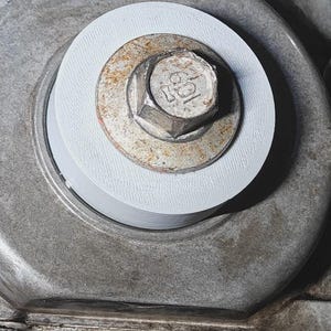 May include: Close-up of an engine component featuring a rusty bolt with a hexagonal head, secured with a white washer. The bolt is centered within a circular metal housing, showcasing mechanical details.