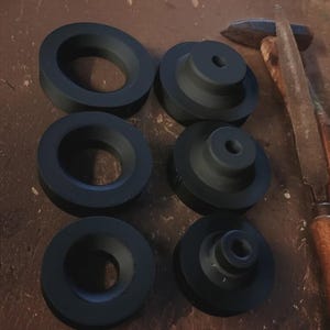 May include: Six black, machined metal parts, three with a central hole and three with a stepped design and a central hole. A hammer and file are in the background, suggesting a workshop setting.