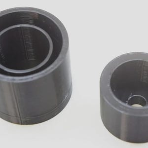 May include: Two dark gray cylindrical objects. One is a taller cylinder with a smaller cylinder inside. The other is a shorter cylinder with a hole in the center. Both have a smooth, matte finish.