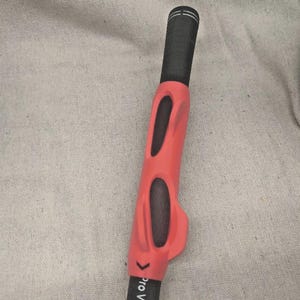 May include: A golf club grip with a black handle and a bright red training aid. The training aid has two oval cutouts and is designed to help with grip. The text "Pro Velvet" is printed on the grip.