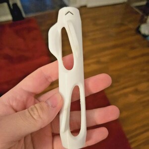Pu&ograve; includere: Un oggetto bianco stampato in 3D con un corpo rettangolare e due grandi ritagli ovali. Un manico curvo si estende dal lato e una piccola forma di freccia si trova nella parte superiore. L'oggetto &egrave; tenuto in una mano.