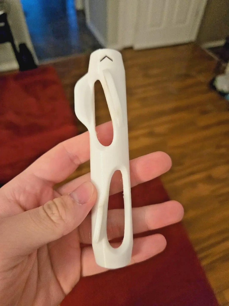 Pu&ograve; includere: Un oggetto bianco stampato in 3D con un corpo rettangolare e due grandi ritagli ovali. Un manico curvo si estende dal lato e una piccola forma di freccia si trova nella parte superiore. L'oggetto &egrave; tenuto in una mano.