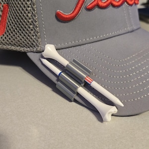 May include: A gray baseball cap with the red embroidered word "Titleist". Two white golf tees with gray holders are placed on the cap. One tee has blue stripes, and the other has red and white stripes.