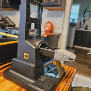 May include: A black and gray knife sharpener with a silver sharpening guide. The sharpener has a base with a yellow logo and a clear plastic guide with measurement markings. The sharpener is on a wooden surface.
