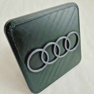 May include: A dark green, rectangular object with a textured surface and rounded corners. The object features a gray Audi logo, consisting of four interlocking rings. The design is likely 3D-printed.