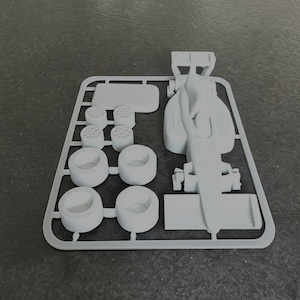 May include: A light gray plastic model kit of a Formula 1 race car. The kit includes the car's body, tires, and other components, all still attached to the sprue. The parts are ready to be assembled.