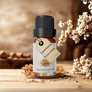 May include: A small amber glass bottle of Frankincense Oil with a black cap and a paper tag. The tag reads "Frankincense Encens d'Inde". The bottle is surrounded by dried frankincense resin and white flowers on a wooden surface.