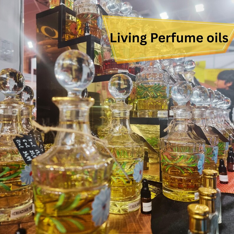 May include: A display of perfume oils in clear glass bottles with decorative stoppers. The bottles are filled with a golden liquid and some have floral designs. The text "Living Perfume oils" is displayed on a yellow banner.