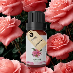 Può includere: Una bottiglia di vetro marrone di olio di rosa con un tappo nero e una piccola etichetta che recita "Rosa Damascena". L'etichetta presenta un'illustrazione di rosa rosa e le parole "Rose Oil". La bottiglia contiene 10 ml di olio, circondata da rose rosa.